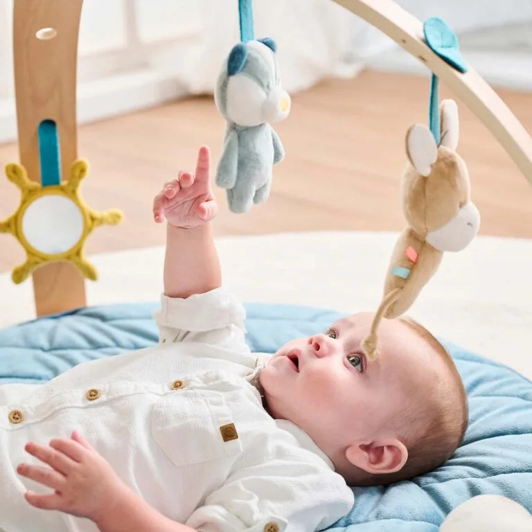 Baby lying on a blue cushion with a wooden crib mobile featuring hanging toys.