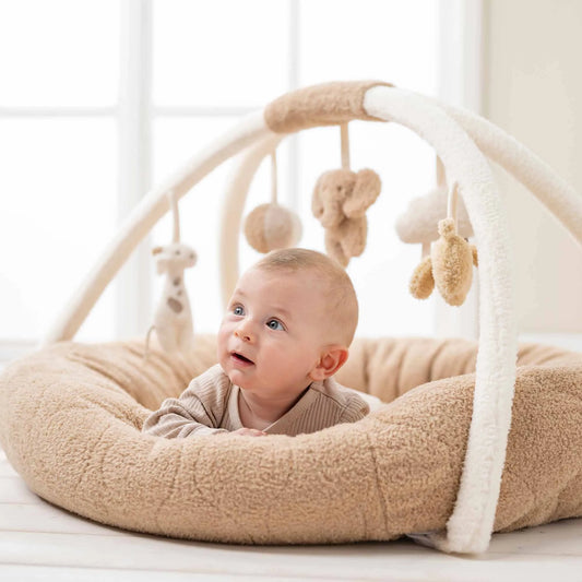 Baby lying on a beige soft play mat with hanging toys in a bright room.