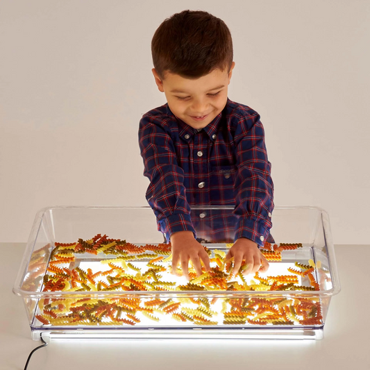 Child playing with dried pasta in tray over light panel
