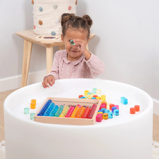 Child playing with colourful acrylic circles toys on a white table in a room with a wooden stool and white wall.