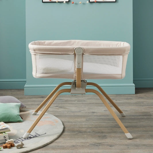 Portable baby crib with beige cover and wooden legs in a room with a teal wall.