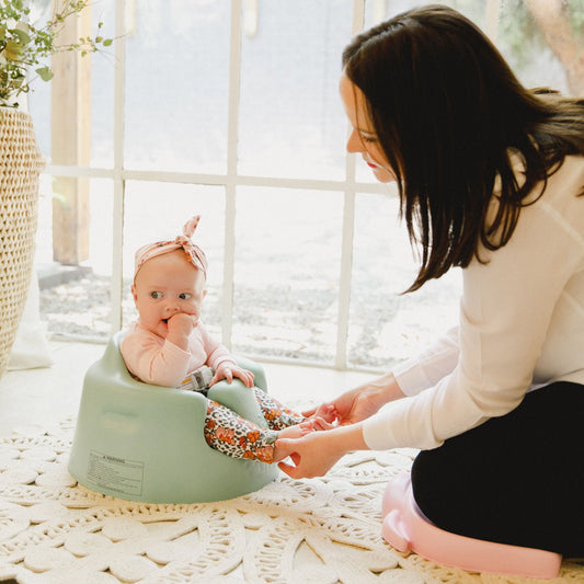 Baby sitting in a green stool with a woman beside them, both on a textured surface with a blurred window background.