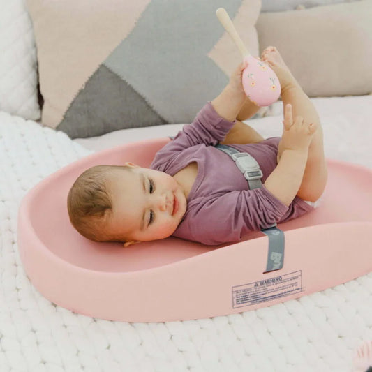 Baby lying on a pink cushion with a toy, surrounded by soft pillows.