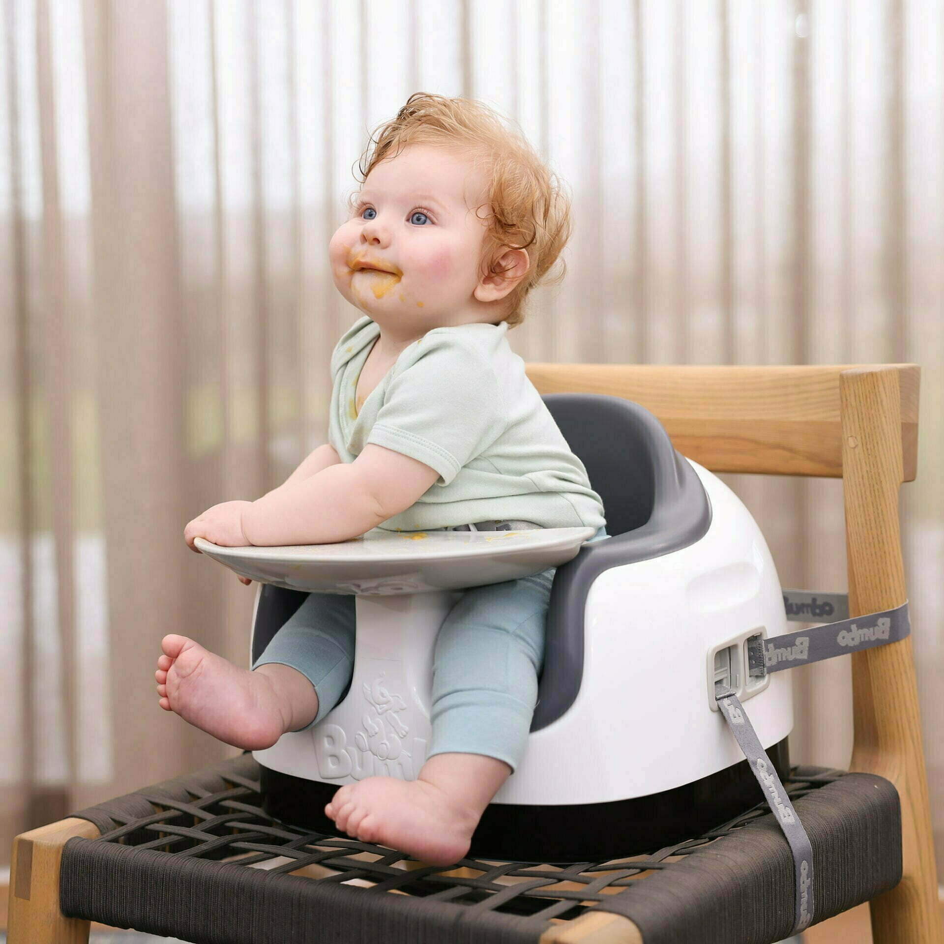 Baby sitting in a high chair with a tray, wearing a light green shirt and blue pants.