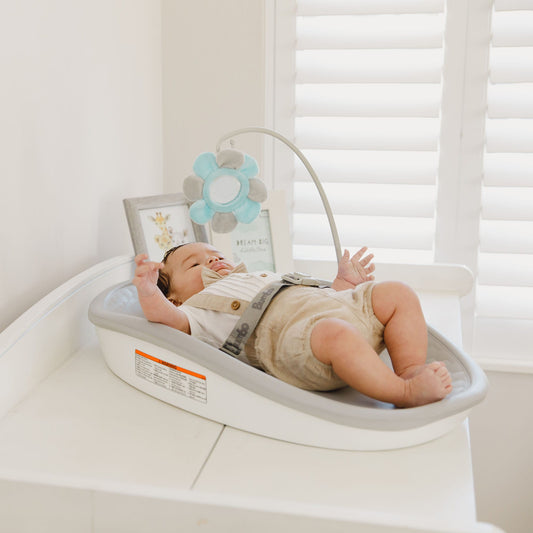 Baby lying on a white baby bouncer with a toy in a room with white walls and window blinds.