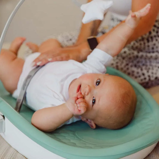 Baby lying in a green baby seat with a person holding a star-shaped toy.