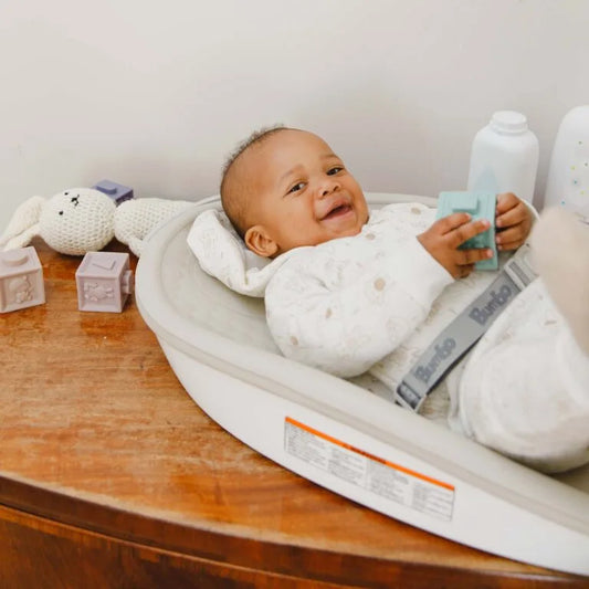 Baby lying on a white cushion with toys and a bottle in the background