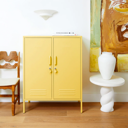 Yellow cabinet in a room with a wooden chair, white vase, and abstract painting.