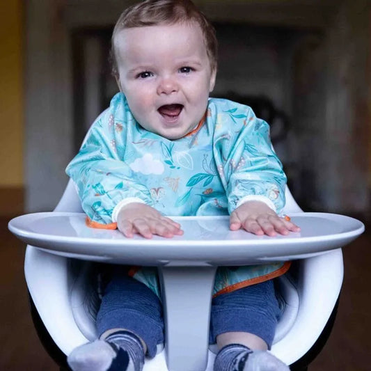 Child in a high chair with a bib, smiling outdoors.