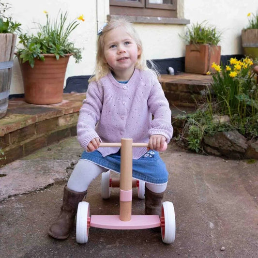 Child sitting on a pink balance bike outdoors with plants and flowers in the background