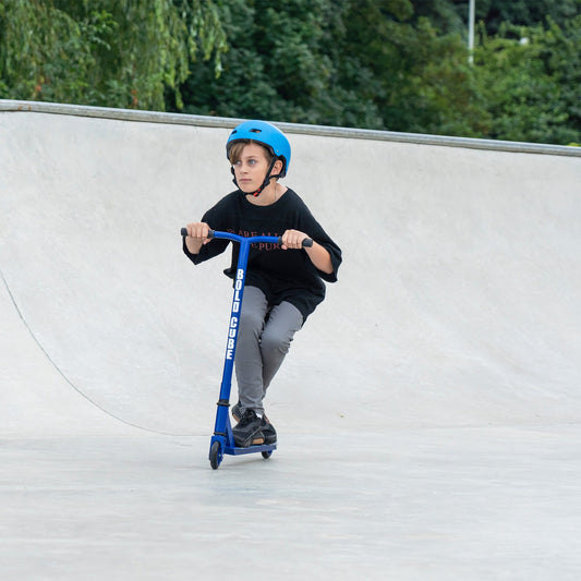 Child riding a blue scooter at a skate park with greenery in the background
