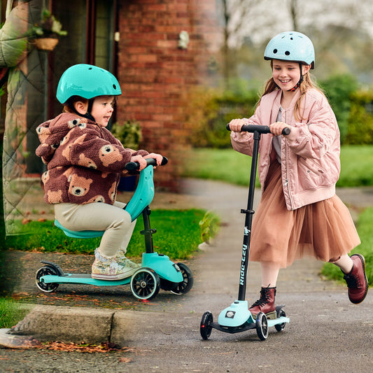 Two children riding scooters on a sidewalk with a brick building in the background