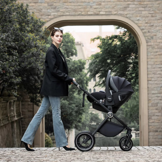 Woman pushing a black stroller in an urban setting with trees and a building in the background.