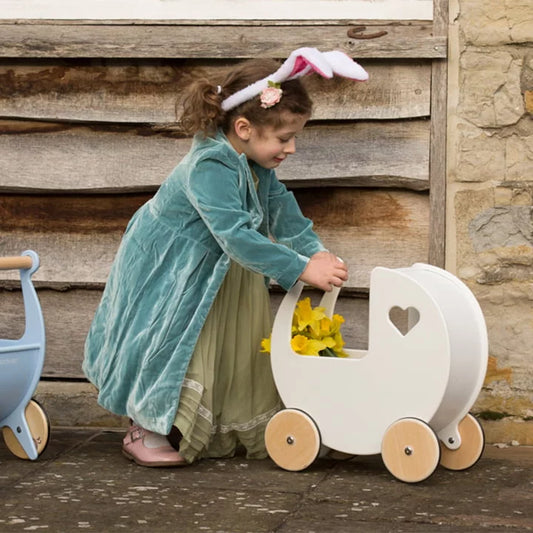 Child in a teal dress with bunny ears pushing a white wooden pram with yellow flowers against a rustic wooden wall.