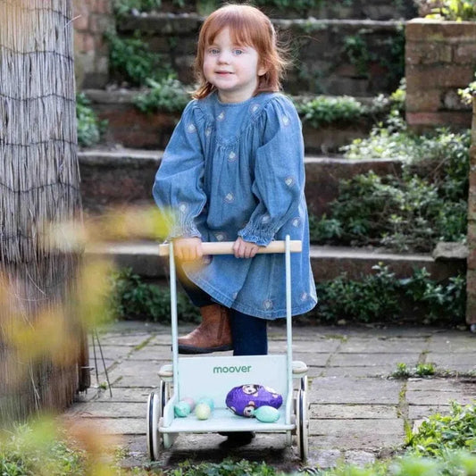 Child in a blue dress pushing a toy cart outdoors on a wooden path.