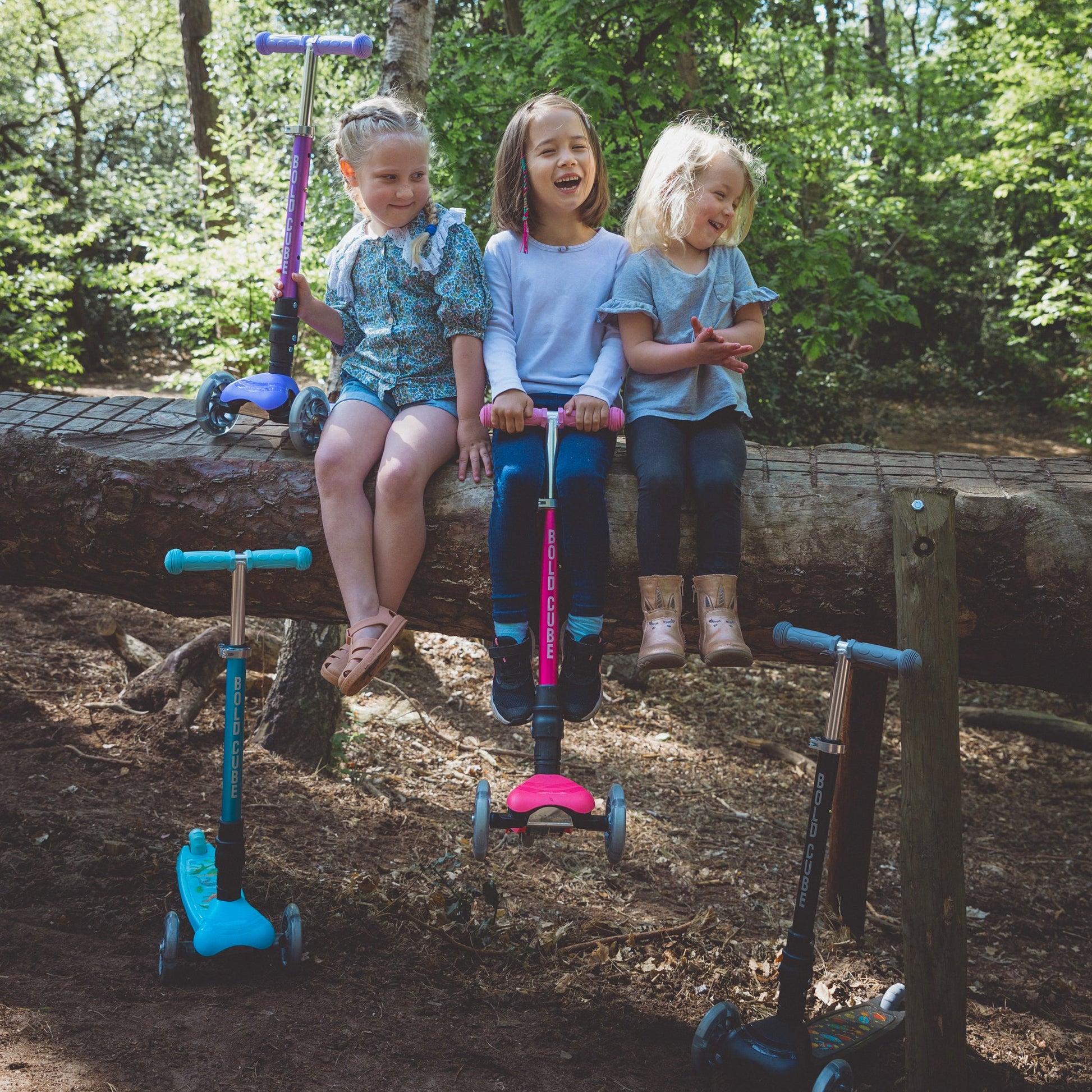 Three children with scooters on a log in a forest setting