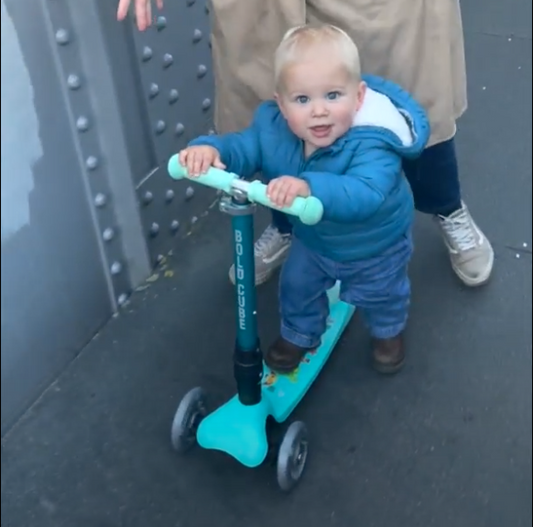 Child in a blue snowsuit riding a turquoise scooter on a sidewalk.