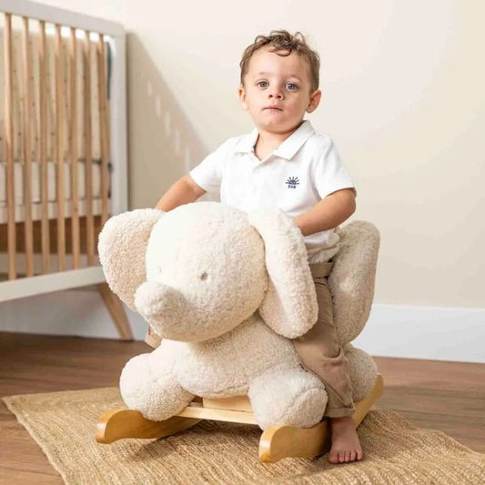 Child sitting on a plush rocking toy in a room with wooden flooring and a crib.