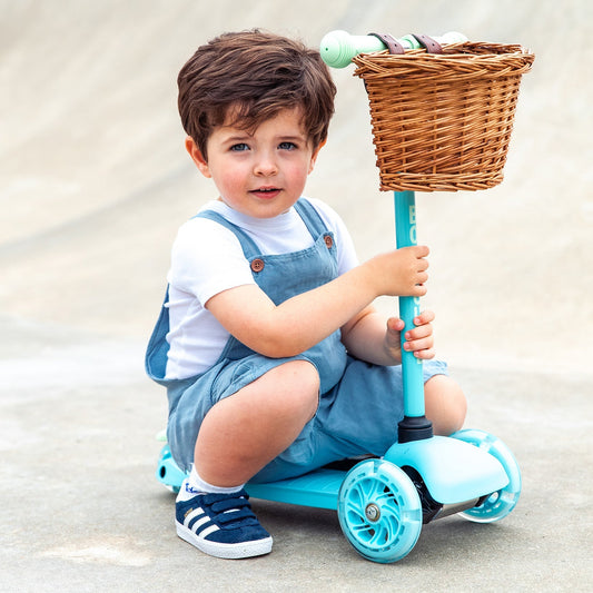 Child sitting on a blue scooter with a wicker basket, outdoors.