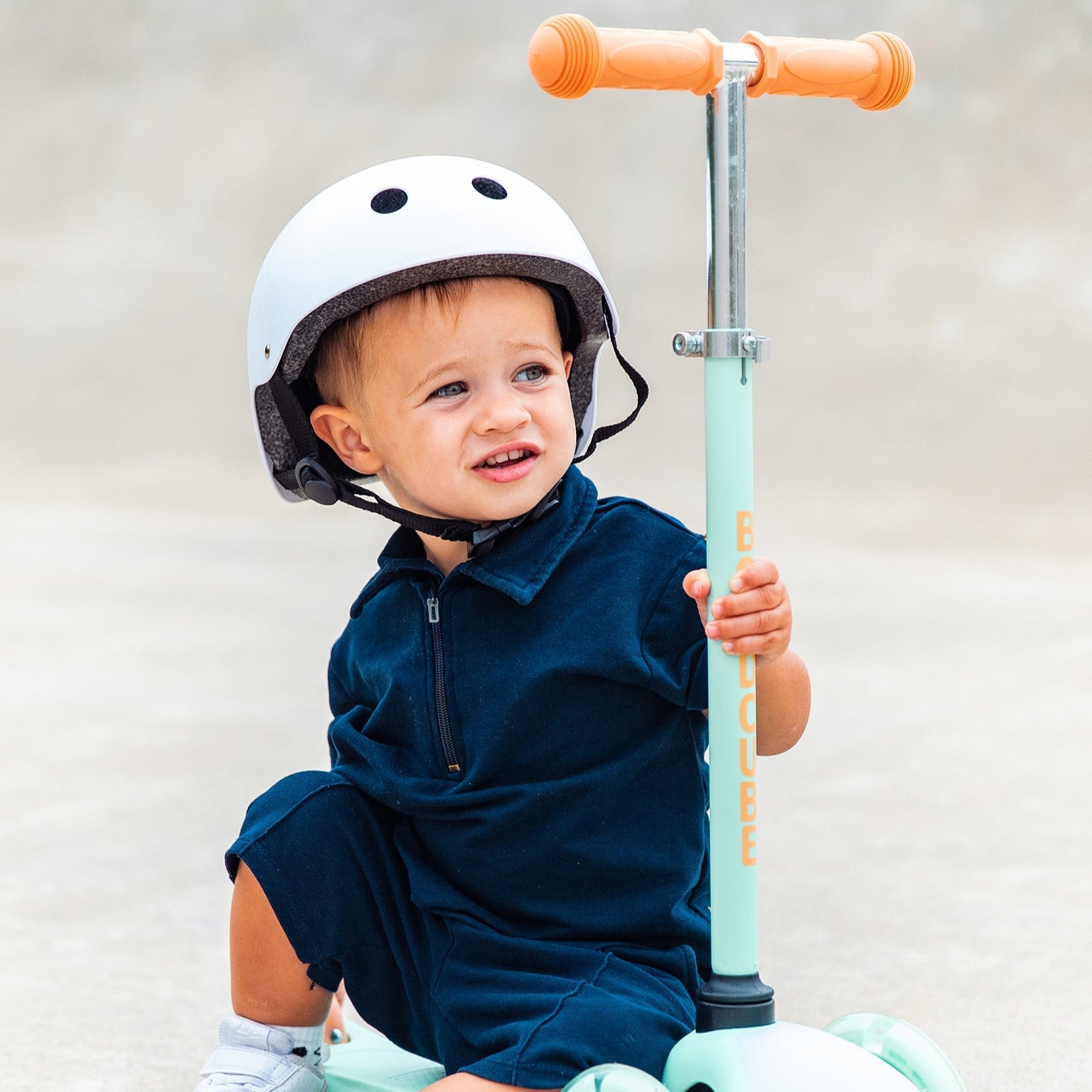 Child sitting on a scooter with a helmet, wearing a navy dress and white shoes.