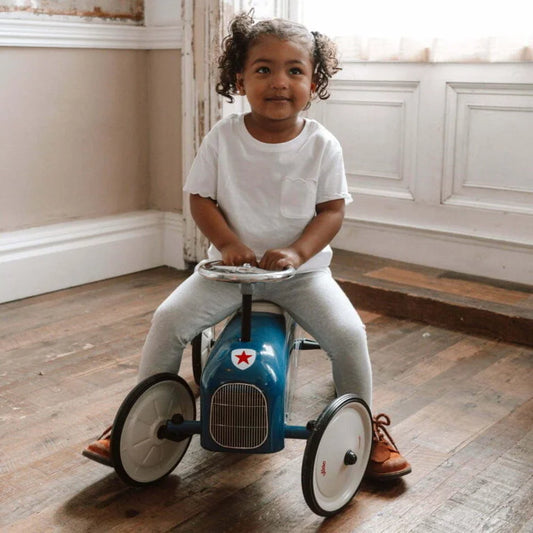 Child sitting on a blue toy car in a room with wooden floor and white walls.