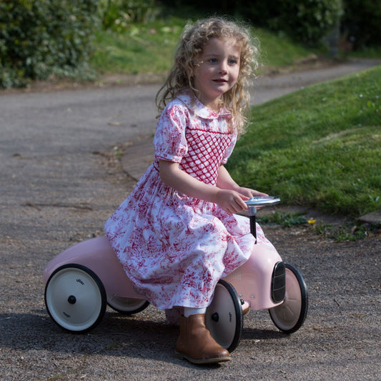 Young girl in a pink and white dress riding a pink toy car outdoors.