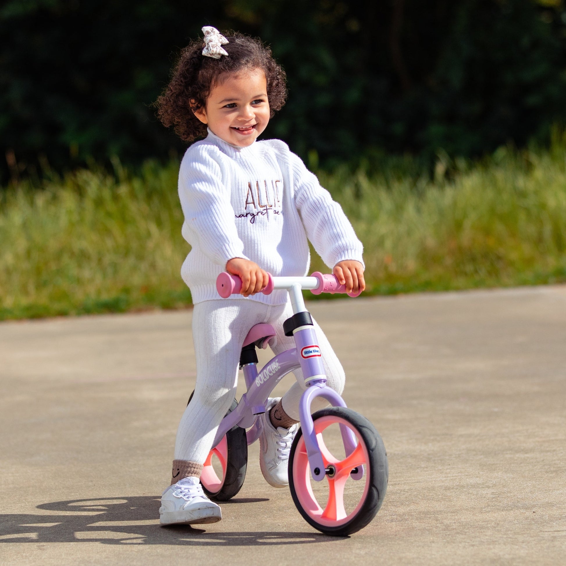 Child riding a balance bike outdoors on a paved path with grass and trees in the background.