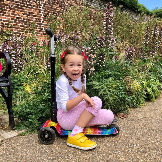 Young girl sitting on a scooter in a garden with flowers and a brick wall in the background