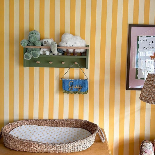 Children's room with yellow and white striped walls, a green shelf with toys, and a wicker basket on the floor.