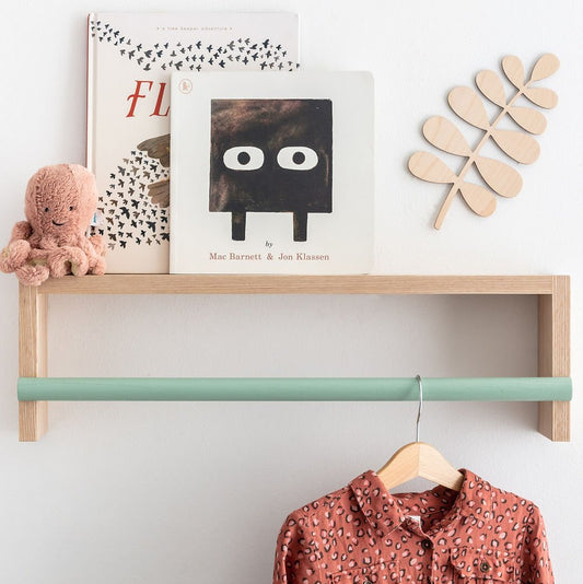 Wooden shelf with books, a teddy bear, and a floral shirt on a hanger against a white wall.