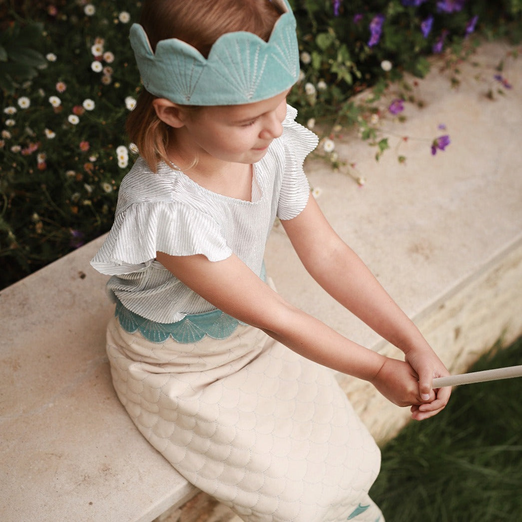 Child wearing a light blue crown and white outfit with ruffled sleeves, sitting outdoors.