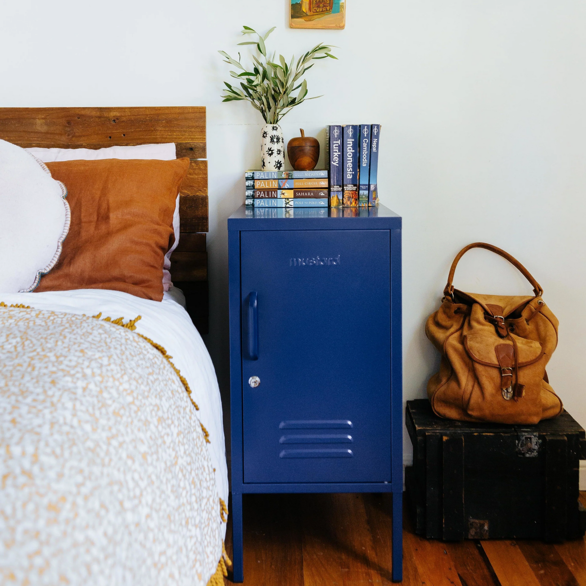 Blue metal locker against a white wall with a bed, plant, and hat in the background.