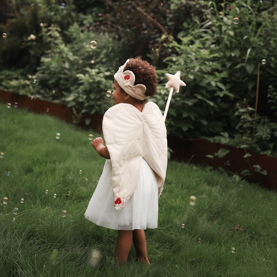 Child in angel costume with wings and star wand standing in a grassy area with trees in the background