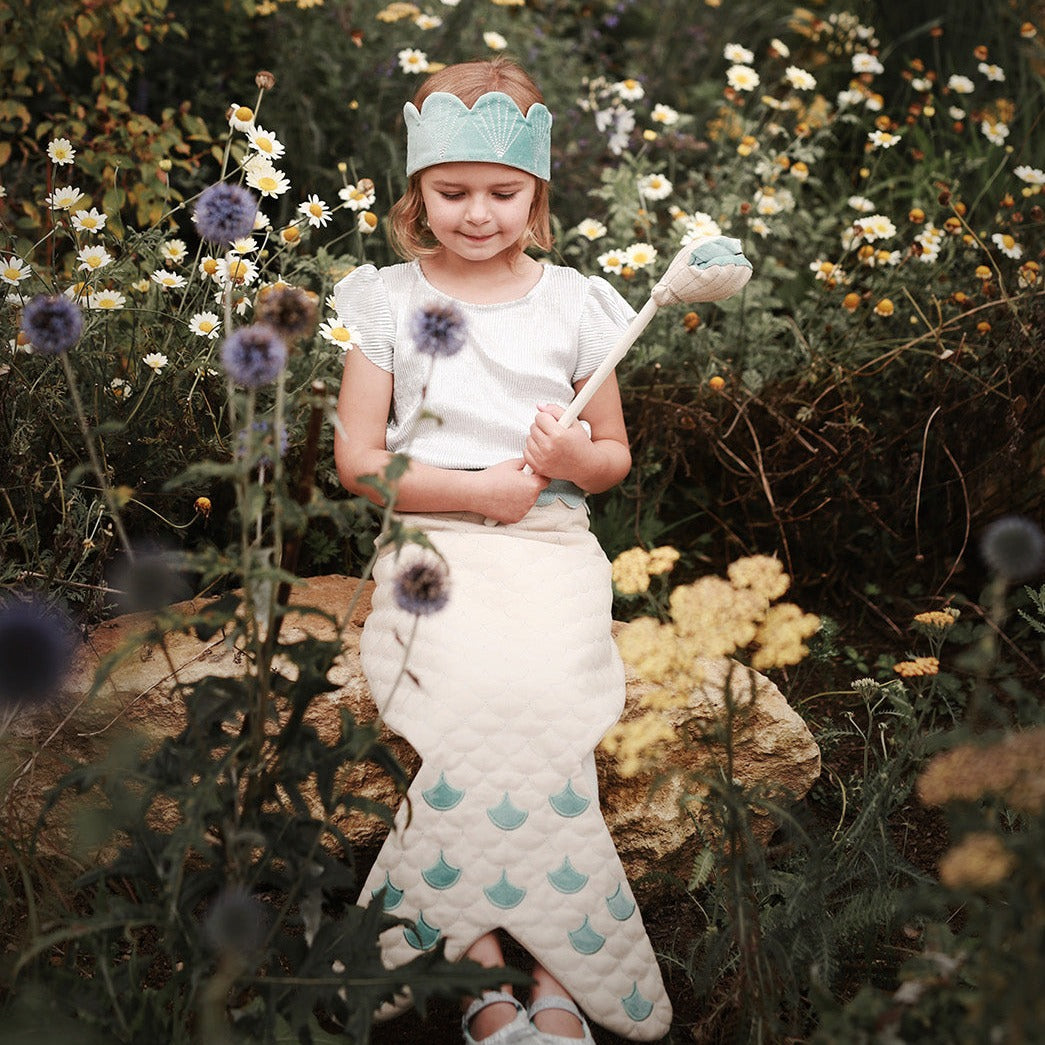 Young girl in a garden with flowers and a broom, wearing a white dress with blue patterns and a headband.