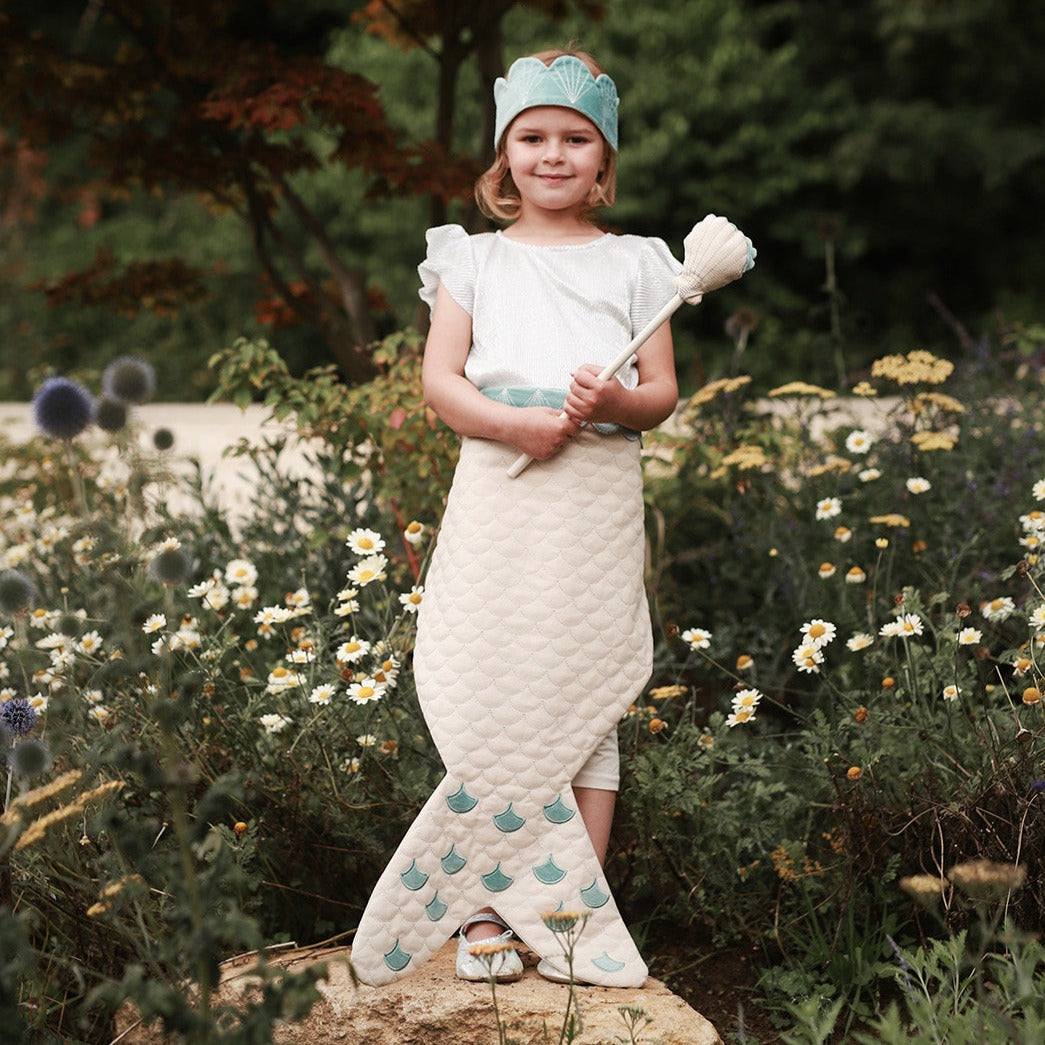 Young girl in a mermaid costume standing in a garden with flowers and plants.