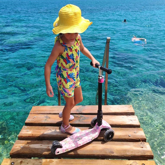 Child with a colorful swimsuit and yellow hat standing on a wooden dock by clear blue water.