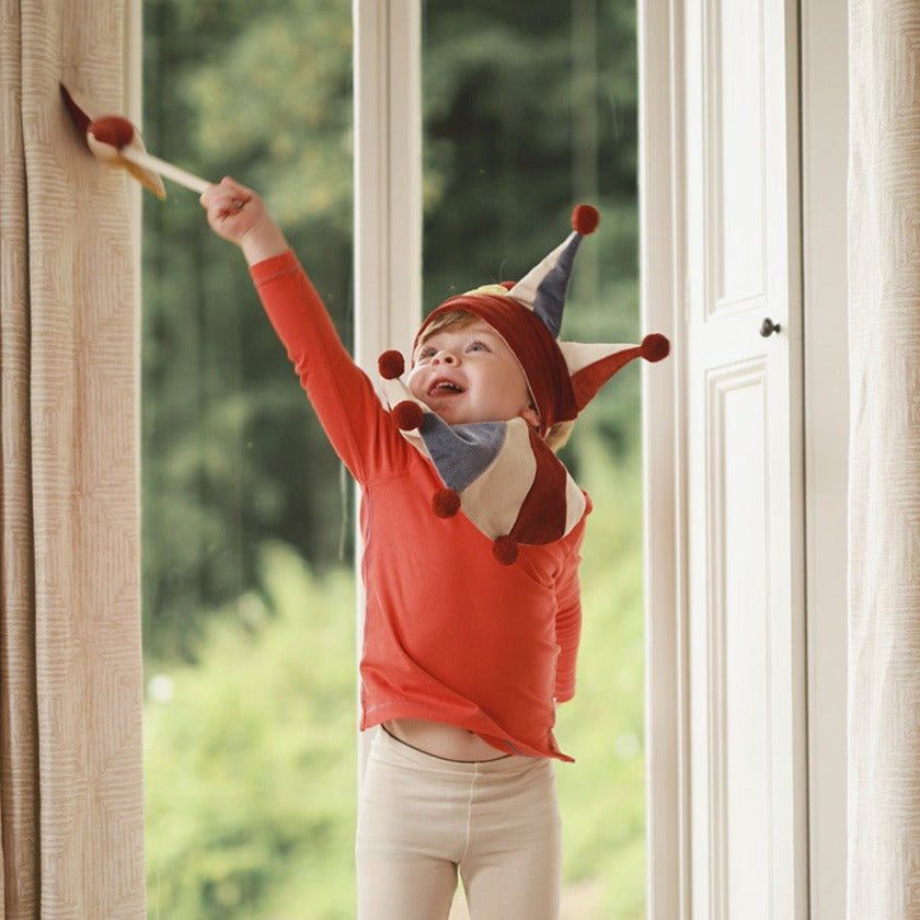 Child wearing a colorful elf hat and red shirt standing by a window with greenery outside.
