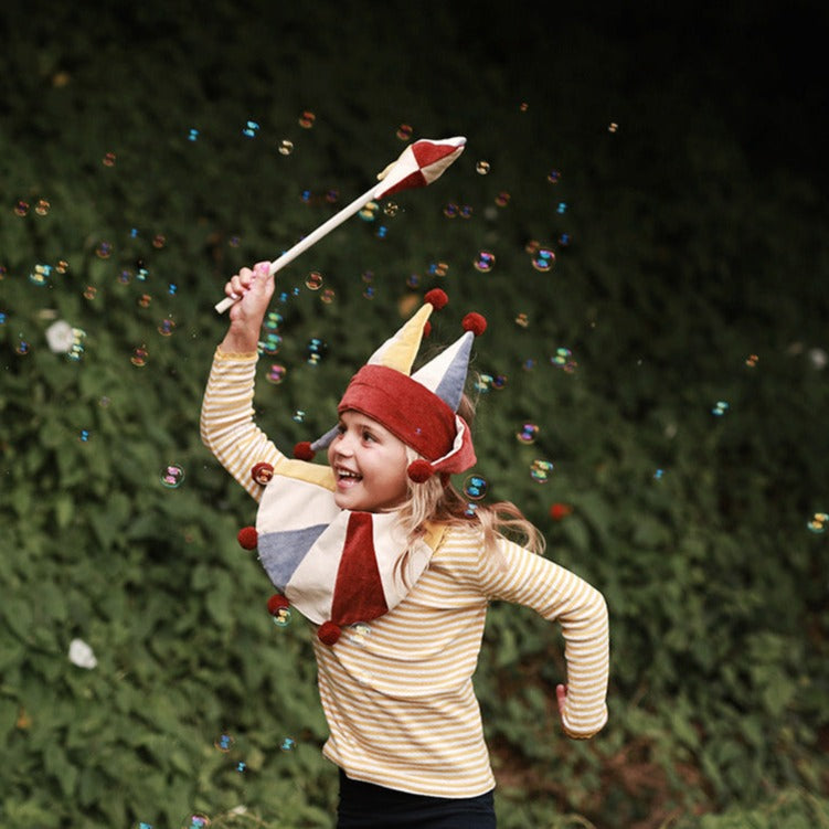 Child wearing a colorful clown hat and striped shirt, holding a wand with confetti against a green background.