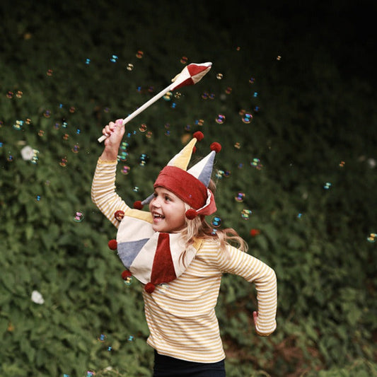 Child wearing a colorful clown hat and striped shirt, holding a wand with confetti against a green background.