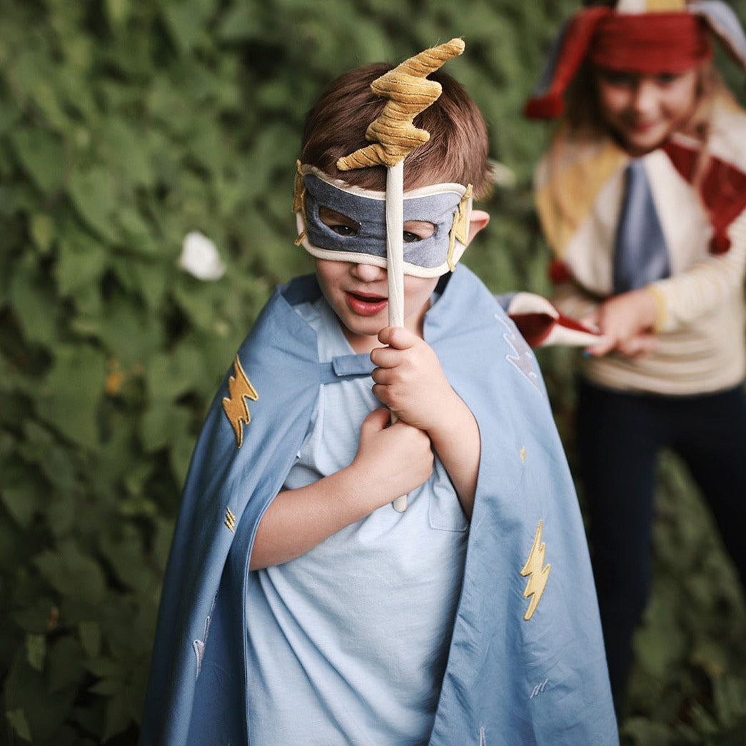 Child in superhero costume with cape and mask, standing outdoors.