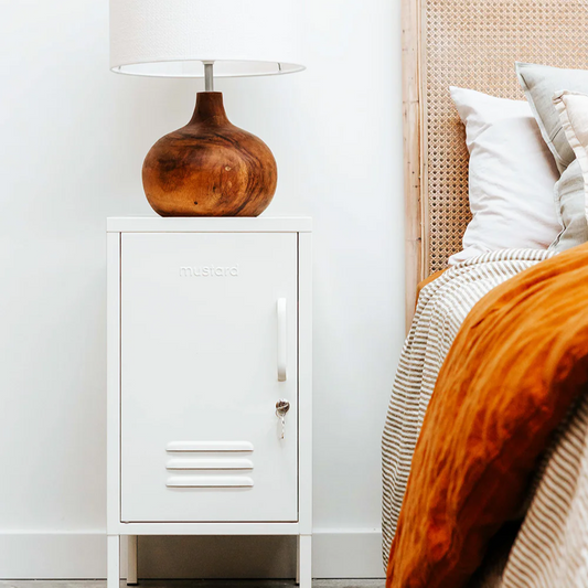 White nightstand with a wooden lamp next to a bed with orange bedding and white pillows.