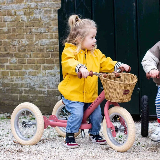 Child in a yellow raincoat riding a pink tricycle with a basket outdoors.