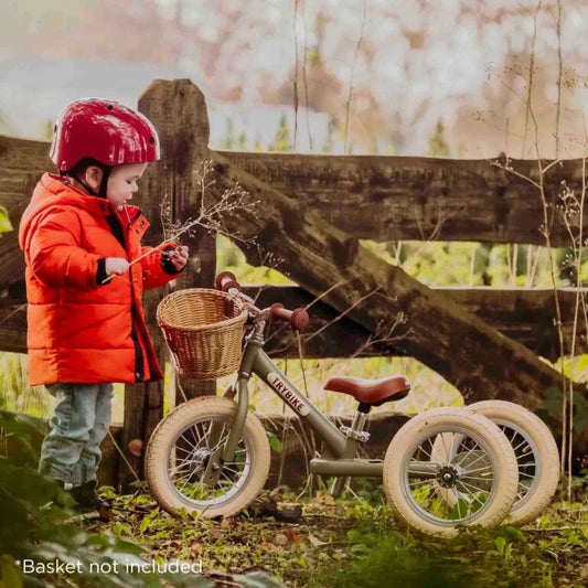 Child in red jacket and helmet standing next to a green balance bike with basket not included, outdoors.