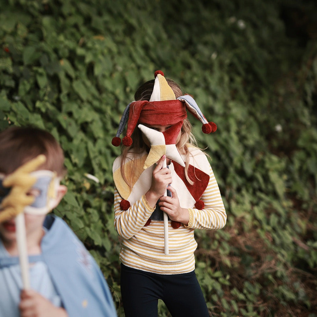 Person wearing a jester hat and mask holding a staff in front of a green bush