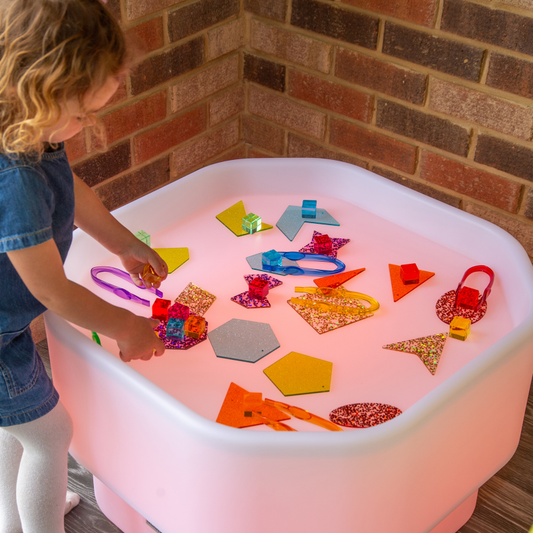 Child playing with colorful geometric shapes in a light-up play table against a brick wall.