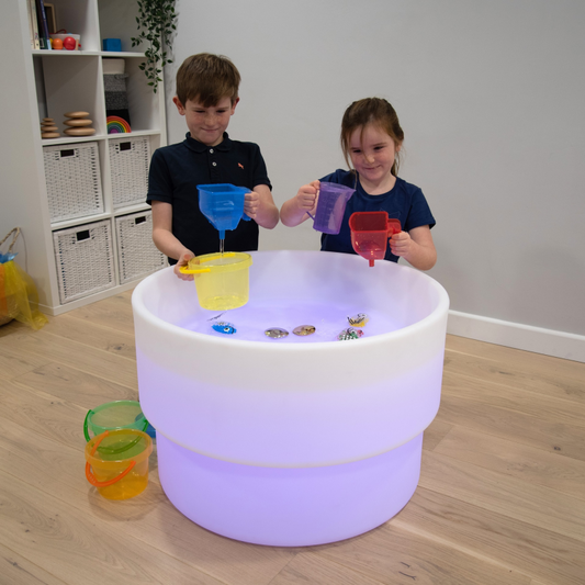 Two children playing with colorful cups over a glowing water table in a room.