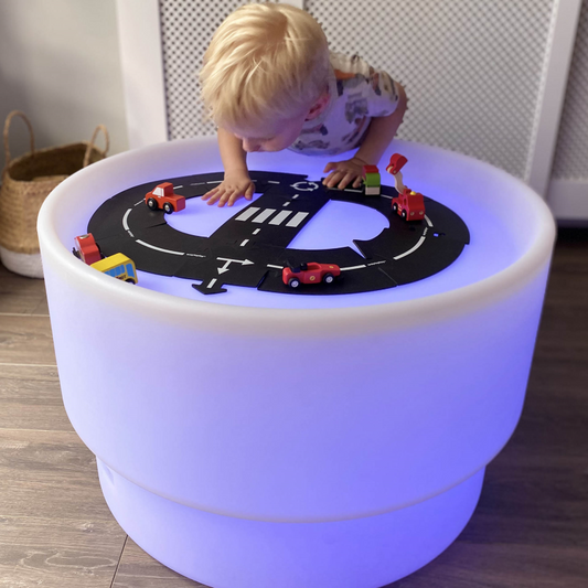 A child standing over the discovery table playing with toy vehicles and road.