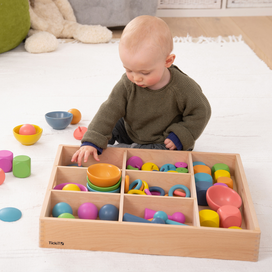 Child playing with colourful toys in a wooden sorting tray on a white floor.