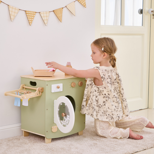 Child playing with a toy washing machine in a room with a window and decorative flags.