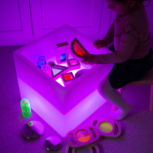 Child playing with a glowing light cube filled with colorful toys in a dimly lit room.